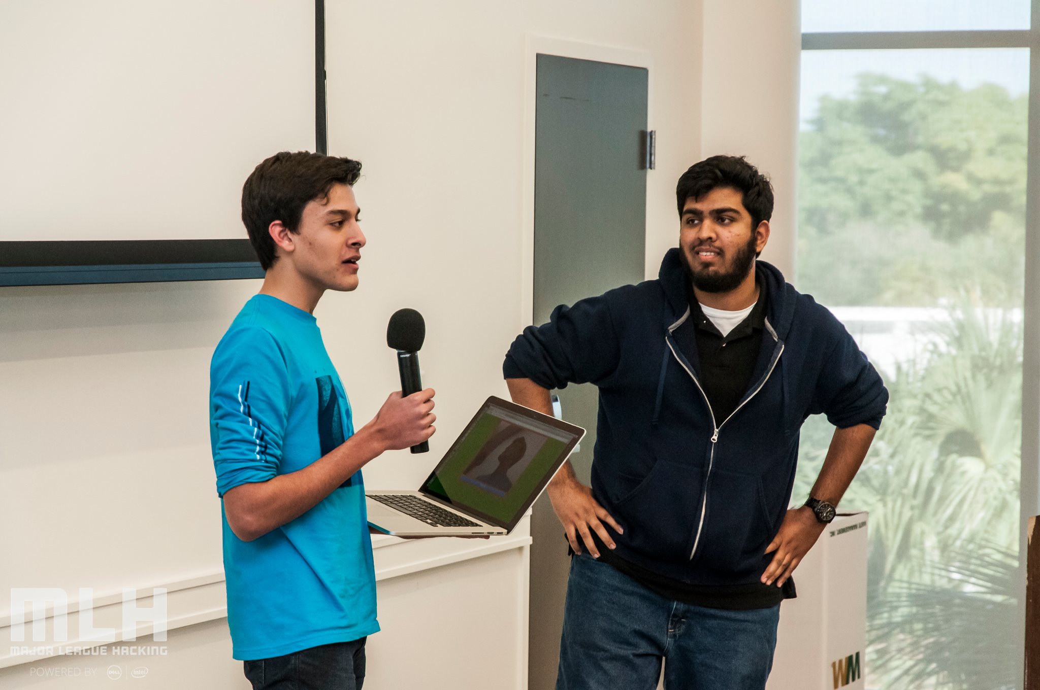 two male students, one holding a laptop and a microphone, presenting their project to the audience